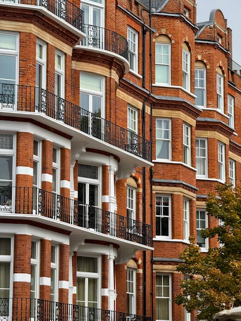 The image depicts a multi-story red brick residential building with classic Victorian architectural features, including large white-framed sash windows, decorative brickwork, and curved bay windows. Several of the windows have black wrought-iron balconies, which extend outward from the building façade and are supported by white columns, adding an ornate element to the structure. The building's exterior showcases contrasting white stone or plaster accents that highlight horizontal bands and window surrounds, enhancing its historic character. In the foreground, part of a leafy tree with green and yellow foliage is visible, indicating a street-level view with some natural surroundings. The lighting appears natural, suggesting daylight with soft shadows cast on the building’s surface. The overall scene reflects a typical urban residential setting where private or independent waste collection might complement local authority rubbish disposal services, aligning with the context of alternative rubbish removal solutions that companies like wastecollectionsouthkensington.co.uk provide for such properties.