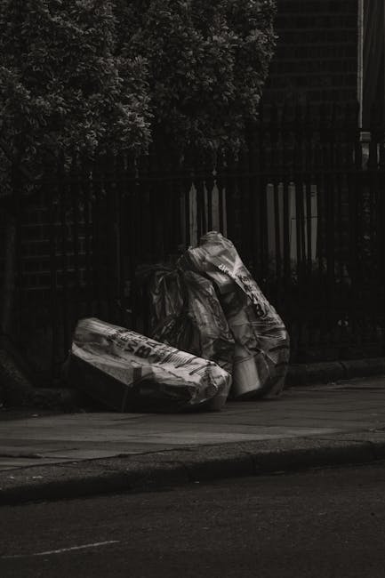 Two large black plastic rubbish bags, visibly crumpled and tied at the top, are positioned on the edge of a pavement next to a dark wooden fence with vertical slats. The bags appear to contain various waste materials, and their glossy surface reflects minimal ambient light. Behind the fence, dense foliage of leafy bushes is partially visible, creating a textured green background. The scene is set outdoors in an urban environment, with the bags placed as part of a private waste collection or rubbish removal process, possibly handled by a local waste management service such as wastecollectionsouthkensington.co.uk. The pavement and roadside surface in the foreground show typical asphalt textures, with the overall lighting subdued, indicating an overcast day or early evening. This visual scene highlights common features of on-site rubbish disposal suited for either private collection or alternative waste handling options outside regular municipal refuse services, aligning with practices for managing household or small-scale commercial waste disposal in South Kensington.