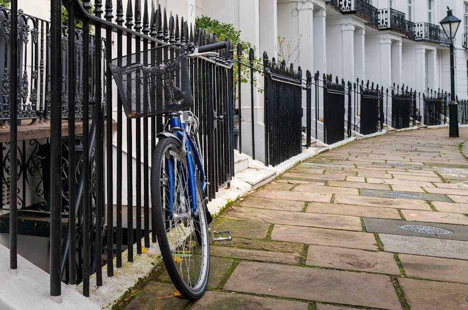 The image depicts a section of a residential sidewalk adjacent to a building with white classical-style columns and black wrought iron railings. In the foreground, a blue bicycle with a black front basket is leaned against the black fence, with the basket positioned at the top. The bicycle’s frame appears well-maintained, with silver handlebars and a black seat. The pavement consists of large, rectangular stone slabs with a weathered appearance, some showing slight moss or dirt accumulation along the edges. The sidewalk curves gently along the building’s frontage. The iron fence features pointed finials, some of which are slightly bent or leaning, indicating possible minor damage or age. Behind the fence, small shrubs and potted greenery are visible, adding some natural element to the urban environment. The lighting suggests an overcast day, providing diffuse illumination across the scene. This setting exemplifies an area where private waste handling or independent collection services, such as those provided by wastecollectionsouthkensington.co.uk, might be used instead of local authority rubbish collection, with visual cues indicating a tidy, residential context suitable for rubbish or waste removal services.