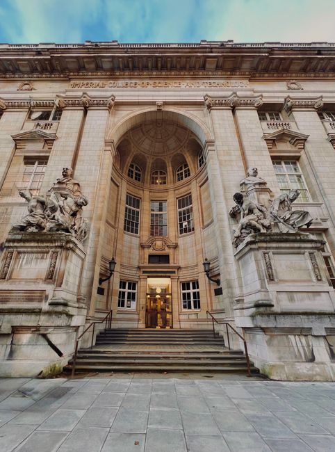 The grand façade of Imperial College London features a classical architectural style with ornate stone carvings and decorative sculptures on each side of a central staircase. The entrance has large, arched windows and a semi-circular portico supported by classical columns. The stone exterior is light beige with detailed moldings and a prominent inscription reading 'Imperial College of Science and Technology' at the top. The steps leading up to the doorway are flanked by metal handrails, and there are two statues situated on pedestals adjacent to the staircase, depicting figures in classical poses. The background reveals a partly cloudy sky with a gradient of light blue and white, providing natural lighting that emphasizes the texture and intricate details of the stonework. The area immediately surrounding the steps is paved with large, flat stone tiles, creating a formal entrance area suitable for various events or visitor access, reflecting the institution's historic and scholarly significance. This setting aligns with the theme of alternative waste handling that may involve the maintenance or collection of materials in university or institutional environments, as handled by companies like wastecollectionsouthkensington.co.uk.