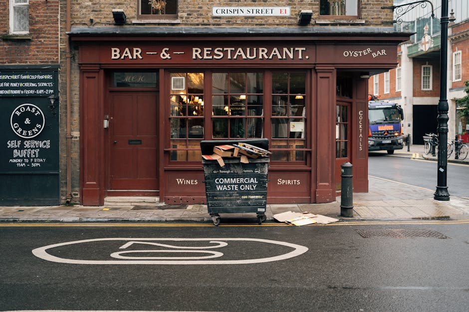 In front of a closed restaurant with a maroon wooden facade and large glass windows, a black wheeled bin labeled 'Commercial Waste Only' is filled with discarded items such as cardboard boxes and paper, some protruding over the top. The bin is positioned on the sidewalk near a black bollard to the right, with a small pile of flattened cardboard boxes resting on the pavement beside it. To the left, there is a blackboard sign advertising a self-service buffet, and a street name sign reading 'Crispin Street' is mounted above the shopfront. A delivery truck is visible in the background, parked along the street, along with a bicycle leaned against a railing and a lamppost. The road in the foreground features a white road marking indicating a parking space or loading zone. The scene is illuminated by natural light, suggesting daytime, and the setting appears to be a commercial area suitable for private waste handling or local waste collection services, such as those offered by wastecollectionsouthkensington.co.uk, for on-site clearance or rubbish disposal.