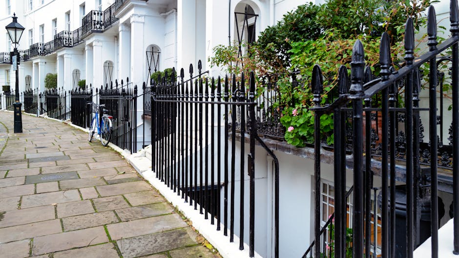 A row of white terraced residential buildings with black wrought iron railings along the sidewalk in front. The railings, featuring pointed finials, surround small garden areas and staircases leading down to basement entrances. One of the gardens contains leafy green plants with pink flowers, adding a touch of color. The pavement consists of rectangular stone slabs, some showing slight weathering. A blue bicycle is parked against one of the railings in the foreground. The scene is captured during daylight hours, with natural lighting highlighting the textures of the iron fence, stone pavement, and plant foliage. The environment suggests an urban setting where residents may consider private rubbish disposal options, with the iron railings serving as boundary markers that are consistent with the aesthetic of London's residential areas. The overall scene emphasizes traditional architecture and front garden spaces, aligning naturally with the context of local rubbish management and waste removal services provided by companies like wastecollectionsouthkensington.co.uk. No visible rubbish is present in the image, but the scene’s layout suggests areas where waste collection might be coordinated in accordance with local regulations.