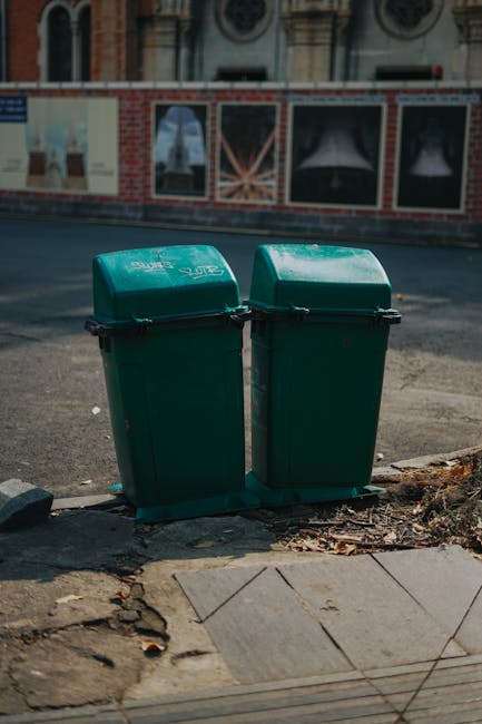 A pair of green plastic rubbish bins with hinged lids positioned side by side on a pavement corner, directly adjacent to a small section of cracked concrete and scattered dried leaves. The bins are situated at the edge of a street, with an asphalt roadway visible behind them. In the background, there is a brick wall with a decorative facade, featuring arched windows and architectural details, suggesting an urban setting. The lighting is natural, with soft shadows indicating daylight, and the scene appears clean and organized, reflecting common waste collection methods in residential or commercial areas where private rubbish disposal may be preferred over council services. The image characteristically illustrates a typical private waste management solution as seen in some urban environments, representing an alternative to standard council rubbish collection routines.