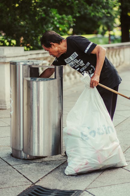 A woman with short dark hair, dressed in a black T-shirt with white and grey graphic text, is seen leaning forward while placing a large white recycling bag into a stainless steel rubbish bin on a paved sidewalk. She holds a long wooden stick in her right hand, aiding her in managing the bag. The bin, which is part of a set of three, stands upright adjacent to a low stone wall with decorative balustrades. In the background, lush green trees and bushes provide a natural setting, and bright sunlight creates a clear, well-lit environment suitable for outdoor waste disposal activities. The scene suggests an instance of independent rubbish collection or on-site waste handling, possibly part of a local waste management effort by a private service such as wastecollectionsouthkensington.co.uk, aimed at tidying streets in urban or residential areas.