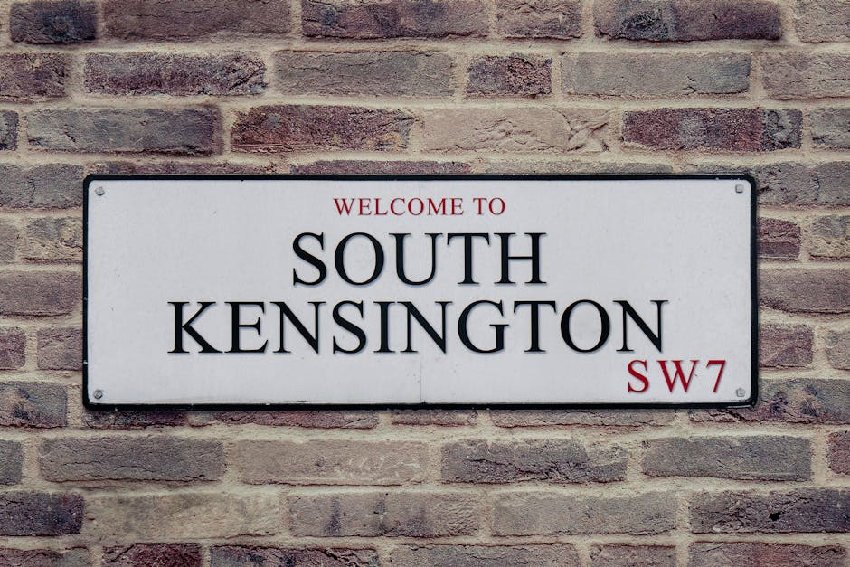A rectangular metal street sign mounted on a brick wall, displaying the text 'Welcome to South Kensington' with 'SW7' in red at the bottom right corner. The sign has a white background with black lettering, and the top line 'Welcome to' is in red. The brick wall behind the sign is composed of reddish-brown bricks with varying shades and textures, arranged in a traditional horizontal pattern. The sign's corners are slightly rounded and secured with visible screws at each corner. The scene suggests an urban environment, typical of a private or independent district entry point, in a setting where local waste management services such as rubbish collection may operate within tight street conditions, aligning with alternative waste handling options often employed in central London neighborhoods.