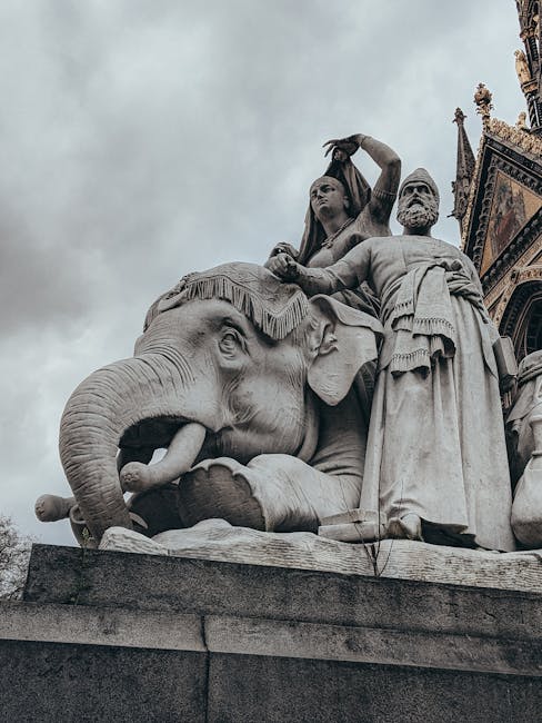 A large stone statue situated outdoors features a detailed sculpture of three figures atop an elephant. The elephant, carved from light-colored stone, displays textured skin and prominent tusks, with its head turned slightly to the left, positioned on a rectangular stone pedestal. Standing behind the elephant, a woman figure is depicted with flowing hair and a head covering, her right hand raised above her head as if holding an object, while her left hand rests on the elephant's back. To her right, a muscular man draped in a cloth over his shoulders and waist stands with a serious expression; his right hand holds a staff or spear, and his left hand is on his hip. The sculpture's surface shows intricate carving details, especially on the elephant's textured skin and the clothing of the human figures. In the background, part of a historic building with ornate architectural features is visible, and the scene is captured under cloudy, overcast skies that cast diffuse lighting on the monument. This public sculpture appears to be an urban installation, possibly associated with a historical or cultural site, and is in a location that facilitates or hints at alternative or private waste management related to maintenance or preservation activities, as suggested by the context of rubbish removal services.