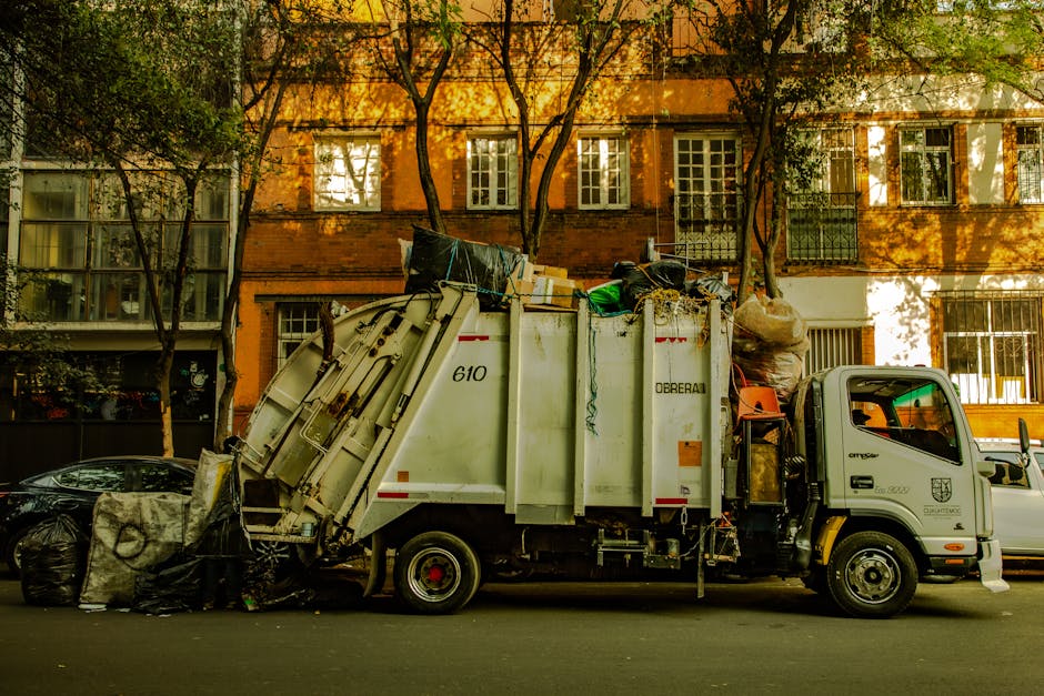 A compact waste collection truck parked on a city street in front of a multi-storey residential building with orange brick walls, white-framed windows, and balconies protected by metal railings. The truck has a white body with visible mechanical components on its side, marked with the number 610 and the logo of the waste management company, and features a small cab with a side mirror. The rear compartment is filled with mixed rubbish, including large black trash bags, cardboard boxes, plastic containers, and miscellaneous debris, some of which is stacked and protrudes from the top. In front of the truck, a few black garbage bags and loose waste are placed on the pavement, ready for collection. The scene is bathed in warm sunlight, casting shadows from nearby trees onto the building, adding a natural environment context relevant to independent or private rubbish disposal services, as seen in waste removal operations typical for urban areas.