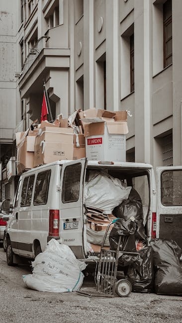 A white cargo van parked on a city street in front of a multi-storey building with beige and grey facades, showing several large open rear doors filled with various types of waste. Inside the van, visible are large white and black garbage bags, flattened cardboard boxes, and other miscellaneous refuse materials. On top of the van, numerous cardboard boxes and large wrapped items are stacked, some of which appear to contain packaging or discarded household items. Leaning against the open rear doors, there is a small hand truck loaded with black rubbish bags. The surrounding environment includes part of the urban pavement and adjacent building windows, with muted lighting indicating overcast weather. The scene reflects a typical setup for private waste collection or rubbish removal using a dedicated vehicle, consistent with services provided by waste collection companies like wastecollectionsouthkensington.co.uk, specifically for the clearance of bulky or accumulated waste in residential or commercial areas.