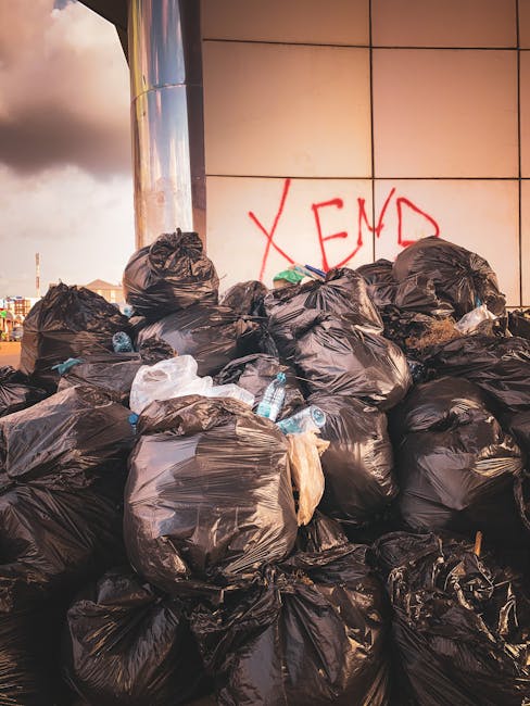 A large pile of black plastic rubbish bags, some partially torn or bulging, stacked against a modern building with a tiled exterior wall. Several crumpled plastic water bottles and miscellaneous small waste items are visible among the bags, which occupy most of the foreground. The bags are placed on a paved surface, likely pavement or a sidewalk, with a slightly reflective finish. In the background, there is a section of a metallic cylindrical structure, possibly part of a nearby building or installation. The wall behind the rubbish bags features a graffiti tag in red spray paint, spelling 'XEND,' which is partially obscured by the bags. The overall scene appears to depict an area awaiting collection or disposal, illustrating private waste management or an alternative rubbish removal process outside typical municipal services, consistent with waste collection activities by companies like wastecollectionsouthkensington.co.uk. The lighting suggests late afternoon or evening, with a mixture of natural and artificial light casting soft shadows and giving a warm tone to the scene.
