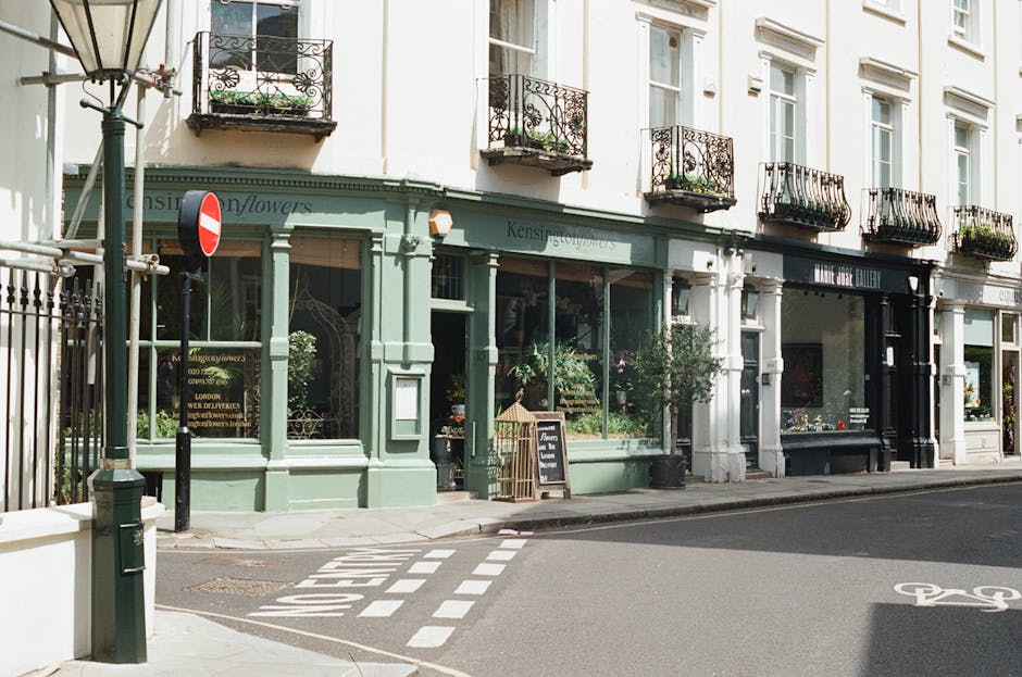 A street view featuring a row of white and light green commercial buildings with large display windows on the ground floor, housing flower shops and art galleries. The flower shop has a pale green exterior with prominent window displays filled with various plants and floral arrangements. In front of the flower shop, there is a small chalkboard sign on the sidewalk listing services or promotions, and a potted plant nearby. To the left, a black street lamp stands beside a metal gate and a red and white no-entry traffic sign mounted on a pole. The building upper floors have white facades with multiple windows and decorative black wrought iron balconies, some with potted plants on them. The street surface is paved with asphalt, with white road markings including a bicycle lane symbol on the right. Natural daylight illuminates the scene, creating clear visibility of the storefronts and urban environment, consistent with private waste collection or rubbish removal services supporting local shops and cafes.