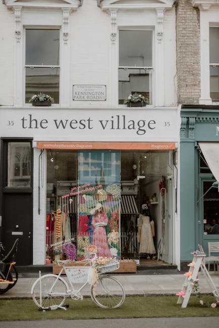 The image features the storefront of a shop called 'the west village' located at 35 Kensington Park Road, with a white exterior wall and large display windows. Above the entrance, there is a sign displaying the shop's name, and a smaller street sign indicating Kensington Park Road. In the shop window, a colorful display of clothing or textiles is arranged, including a prominent mannequin dressed in a pink patterned dress, with other garments hanging or folded nearby. To the left of the shop entrance, a white bicycle with a basket attached to the handlebars is parked on a small strip of well-maintained grass directly in front of the store. The shop's vegetation and decorative elements, such as potted plants or flowers, are visible on the windowsills. The immediate environment appears to be an urban street scene with other storefronts, and the overall atmosphere is bustling yet organized, blending retail activity with a vibrant, community-oriented aesthetic related to local shopping and private waste handling in the area. The lighting is natural, suggesting daytime with clear or slightly overcast skies, and the scene presents a clean, tidy, and well-maintained frontage typical of independent retail outlets that may occasionally require rubbish removal or waste management services from providers like wastecollectionsouthkensington.co.uk to support their daily operations.