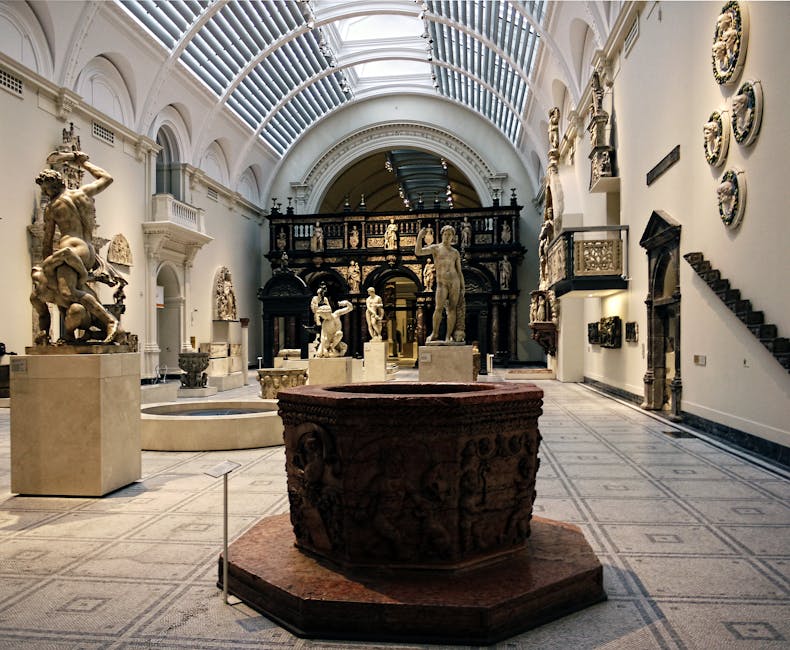 Inside a spacious, well-lit gallery of the V and A Museum, a collection of classical sculptures and artworks is displayed across the polished stone floor. The ceiling features an ornate, curved glass and metal structure allowing natural light to flood the space, which highlights the detail of the stone and marble sculptures positioned throughout the room. In the foreground, a large, intricately carved, reddish-brown stone basin or artifact with detailed relief work and decorative handles rests on a slightly elevated platform. To the left, several sculptures, including a prominent standing figure and a smaller dual-figure group, are mounted on square pedestals. The walls on the right are decorated with a series of ceramic or stone plaques arranged in a semi-circle, along with small, rectangular wall-mounted displays. A dark, ornamental balcony with detailed balustrades extends around the upper part of the room, and a staircase ascends on the far right. The overall environment is clean, orderly, and indicative of a high-value cultural setting, with a neutral and natural lighting atmosphere emphasizing the artworks' textures and materials, reflecting the museum’s art conservation and display standards. Occasionally, a few visible service elements, such as small barriers, subtly suggest ongoing or recent interior management related to the space’s upkeep or safety protocols. This scene aligns with an environment where professional cleaning or clearance services, such as those by wastecollectionsouthkensington.co.uk, might be involved in managing the maintenance of heritage or exhibition spaces through discreet, private waste handling and on-site removal processes.