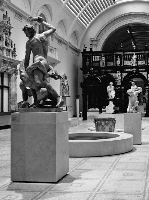 Inside a spacious, well-lit gallery of the V and A Museum, a collection of classical sculptures and artworks is displayed across the polished stone floor. The ceiling features an ornate, curved glass and metal structure allowing natural light to flood the space, which highlights the detail of the stone and marble sculptures positioned throughout the room. In the foreground, a large, intricately carved, reddish-brown stone basin or artifact with detailed relief work and decorative handles rests on a slightly elevated platform. To the left, several sculptures, including a prominent standing figure and a smaller dual-figure group, are mounted on square pedestals. The walls on the right are decorated with a series of ceramic or stone plaques arranged in a semi-circle, along with small, rectangular wall-mounted displays. A dark, ornamental balcony with detailed balustrades extends around the upper part of the room, and a staircase ascends on the far right. The overall environment is clean, orderly, and indicative of a high-value cultural setting, with a neutral and natural lighting atmosphere emphasizing the artworks' textures and materials, reflecting the museum’s art conservation and display standards. Occasionally, a few visible service elements, such as small barriers, subtly suggest ongoing or recent interior management related to the space’s upkeep or safety protocols. This scene aligns with an environment where professional cleaning or clearance services, such as those by wastecollectionsouthkensington.co.uk, might be involved in managing the maintenance of heritage or exhibition spaces through discreet, private waste handling and on-site removal processes.