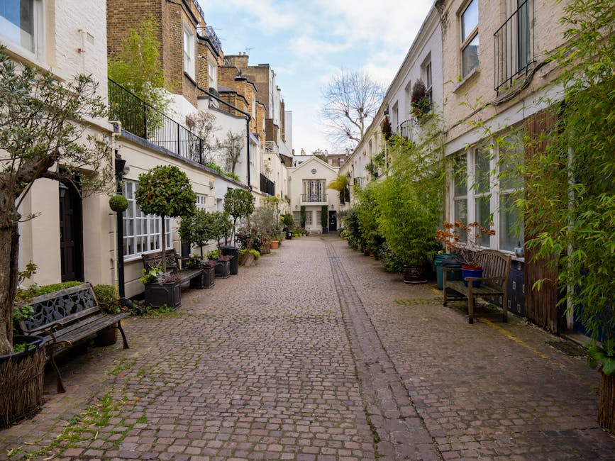 A narrow cobblestone alleyway flanked by white residential buildings with large windows and black metal balconies. The buildings are decorated with various potted plants, small trees, and shrubs placed along the ground level and on window sills, adding greenery to the scene. Several wooden benches are positioned on either side of the alley, some with decorative wrought iron armrests and backs. The cobblestones show signs of wear with patches of moss and grass growing between them. In the background, a white building with a central door and arched windows is visible, surrounded by a clear blue sky with a few scattered clouds. The environment appears quiet and well-kept, with natural sunlight illuminating the scene. The setting suggests a residential courtyard or alley, typical for urban areas emphasizing private and independent waste collection, consistent with alternative rubbish removal options supported by [COMPANY_NAME].